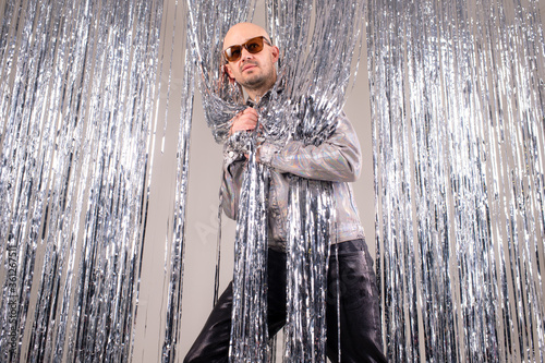 Strange caucasian man in a glittering white shirt, black leather pants, grey shoes and black sunglasses fooling around shiny silver tinsel photo