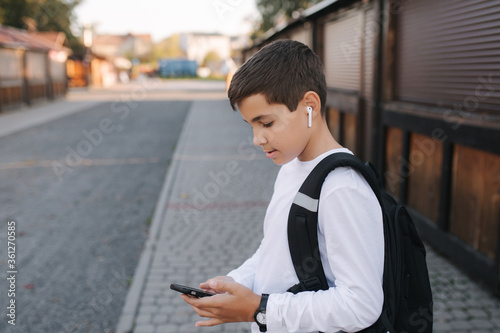 Teenage boy in white sweatshirt and with black backpuck use phone and wireless headphones and print message in social network
