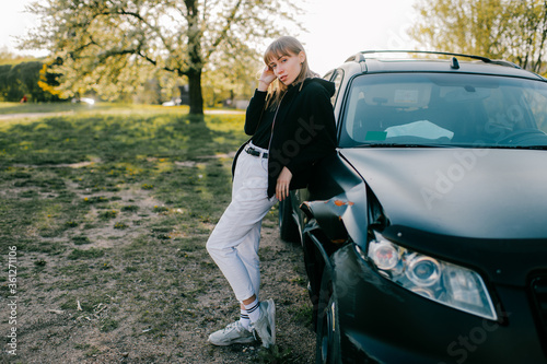 Beautiful young caucasian woman in white jeans, white trainers and black jacket take photos with black broken car