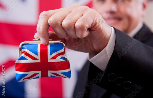 Stereotypical English businessman holding a Union Jack money purse. The man is wearing a dark business suit and bowler hat, with a Union Jack flag background.