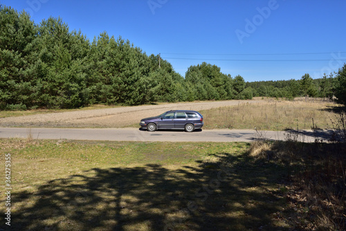 Samochód kombi w ciemnym kolorze na asfaltowej drodze wśród drzew w letni dzień. 