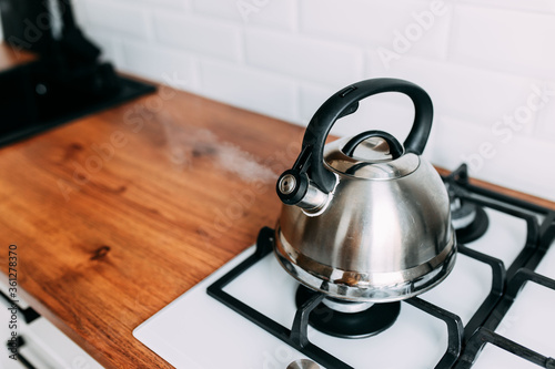 The kettle boils, steam from metal teapot on gas oven. Bright kitchen interior. White modern dining room. Wooden complete kitchen with gas oven. photo