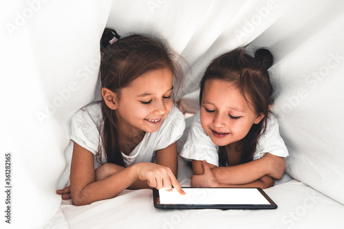 Two beautiful little sisters lying in the bed and look at the screen of a tablet, smart technology