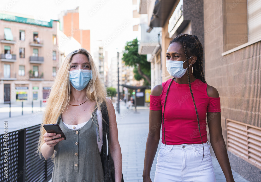 Two women of different ethnicity walking on street with a protective mask