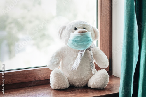 Covid-19 stay at home for children quarantine, illness concept. White fluffy teddy bear wearing blue medical face mask sitting on window