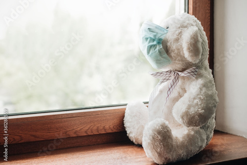White teddy bear wearing blue medical face mask sitting on windowsill looking through the window. Covid-19 pandemic, quarantine, social distancing, stay at home concept.