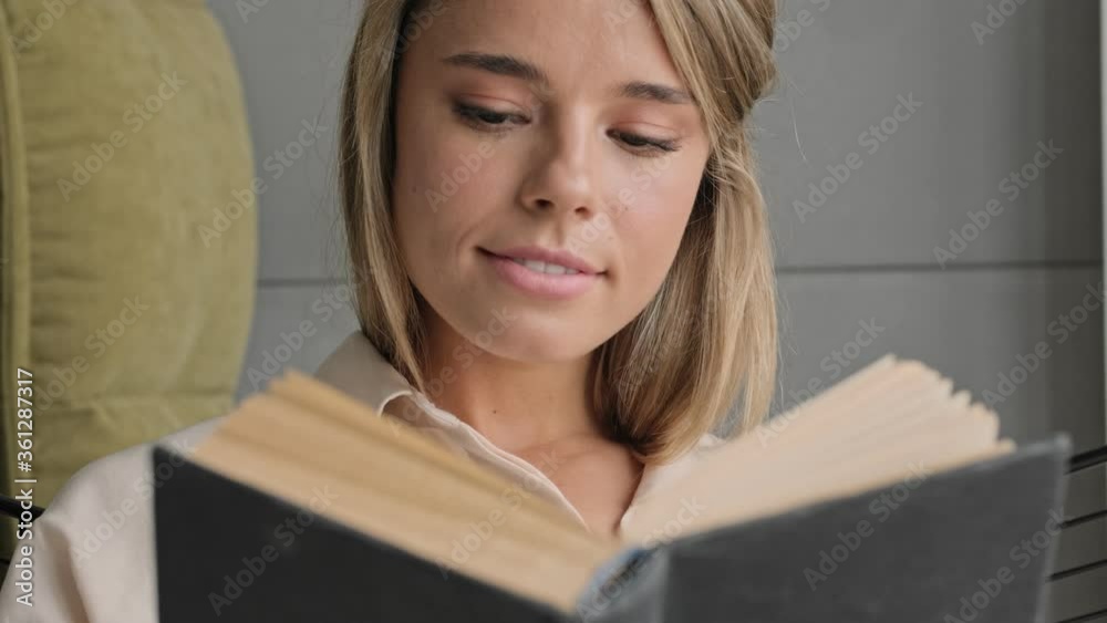 Smiling attractive blonde woman reading book while sitting on chair at home Smiling attractive blonde woman reading book while sitting on chair at home