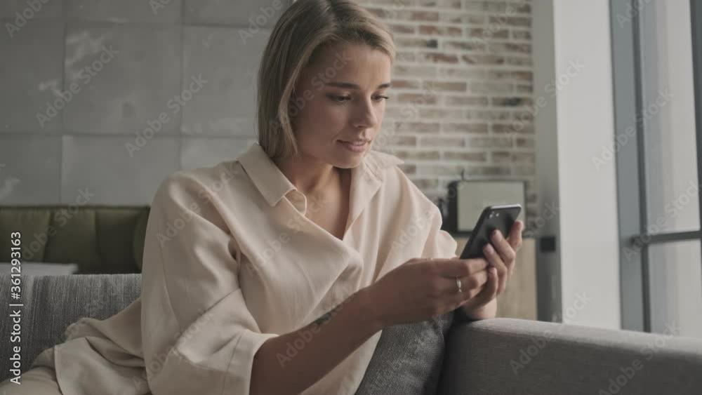 Smiling attractive blonde woman using smartphone while sitting on sofa at home Smiling attractive blonde woman using smartphone while sitting on sofa at home