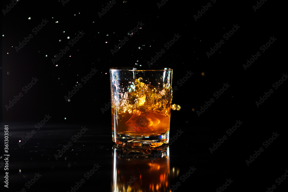 Ice cube falls into a glass with a whiskey on black background. Fluid in motion. Droplets of spray Ice cube falls into a glass with a whiskey on black background. Fluid in motion. Droplets of spray