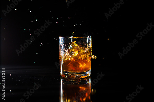 Ice cube falls into a glass with a whiskey on black background. Fluid in motion. Droplets of spray