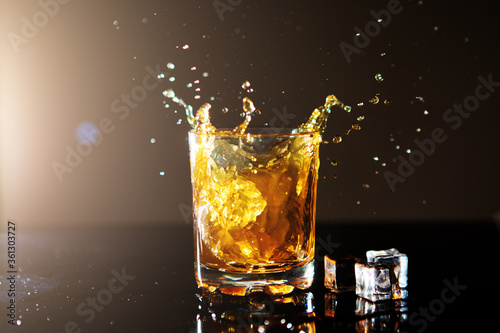 Glass of whiskey with ice cubes on black background. Spray of whiskey.