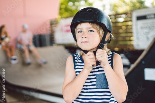 Portrait of handsome caucasian boy athlete skateboarder in protective helmet with skateboard in hands looking at camera on background of skate park. A child and an active hobby, sports and health © Elizaveta