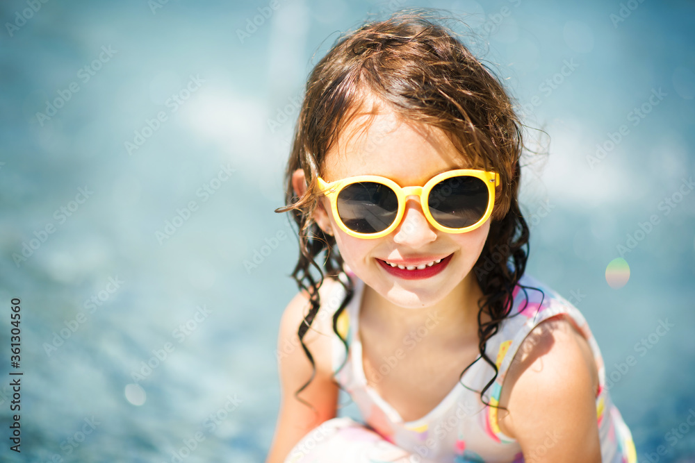 Close-up portrait of cheerful little smiling girl in summer yellow sunglasses with blue water on background. Summer vacation at sea. Happy child in summer hot day. Copy space for text