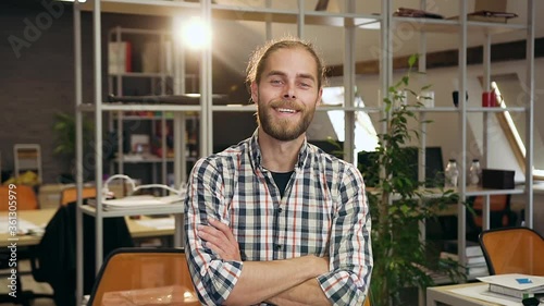 Good-looking satisfied professional confident young male worker with beard looking at camera with crossed arms in modern workroom photo