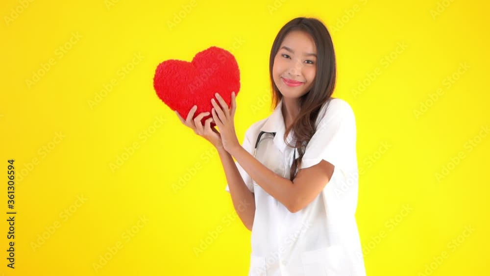 Pretty asian female doctor showing red heart symbol, slow motion, isolated on yellow background Pretty asian female doctor showing red heart symbol, slow motion, isolated on yellow background