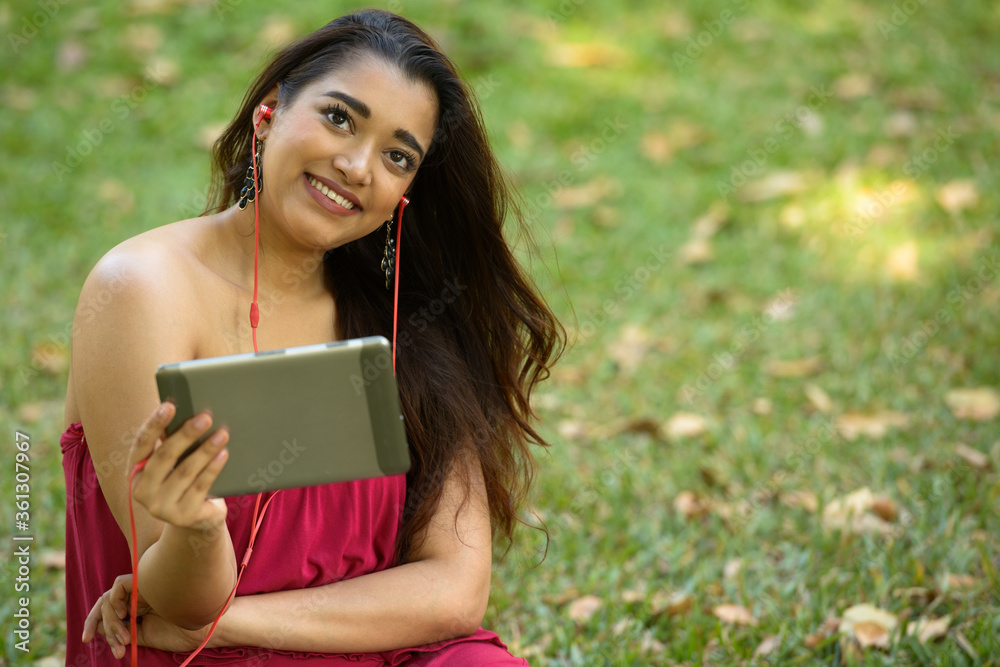Happy young beautiful Indian woman thinking while using digital tablet at the park Happy young beautiful Indian woman thinking while using digital tablet at the park