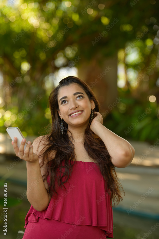 Happy young beautiful Indian woman thinking while using phone at the park Happy young beautiful Indian woman thinking while using phone at the park