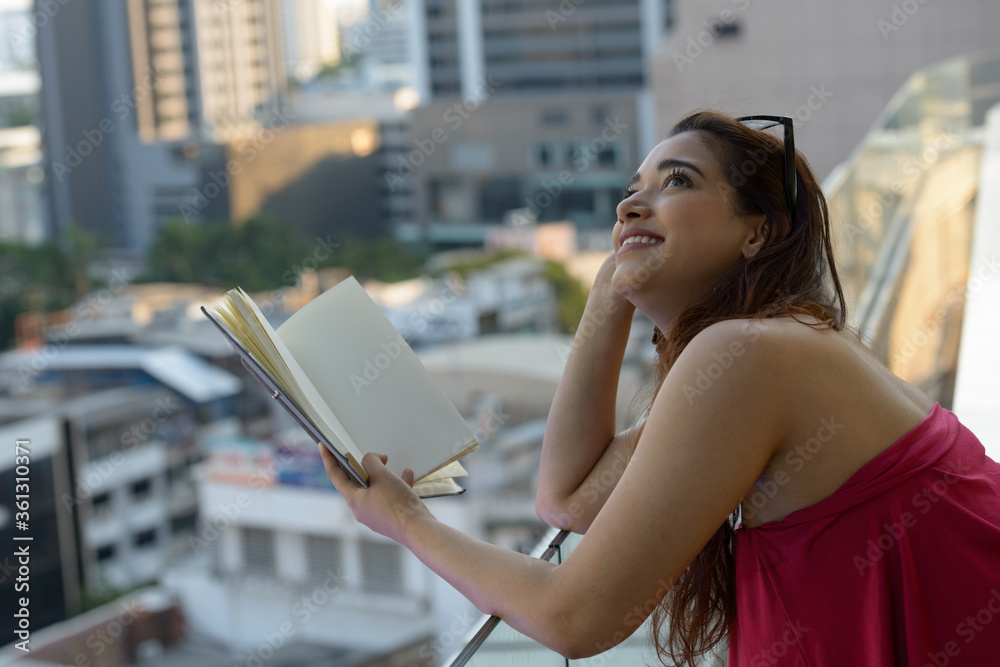 Fényképezés Happy young beautiful Indian woman thinking while reading book against view of t