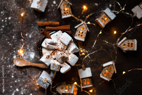 Winter hot drink: hot chocolate with marshmallows, cinnamon , anise. Christmas lights in the shape of houses. Decorative snow, dark wooden background. Festive mood, cozy atmosphere. Flat lay top view