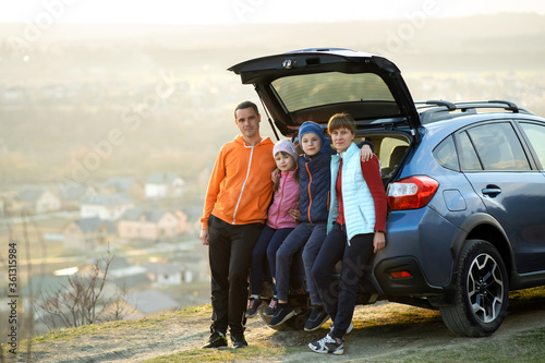 Happy family standing together near a car with open trunk enjoying view of rural landscape nature. Parents and their kids leaning on vehicle luggage compartment. Weekend travel and holidays concept. © bilanol