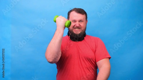 fat fancy man in a pink t-shirt with dumbbells funny doing exercises on a blue background.