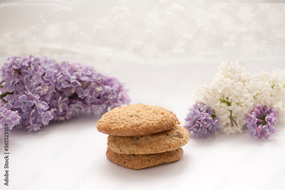 Three oatmeal cookies. In the background, lilac and white lilac flowers. White background Three oatmeal cookies. In the background, lilac and white lilac flowers. White background
