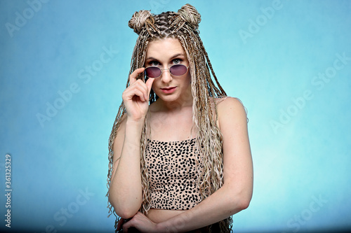 Portrait of a caucasian fashionable woman in glasses with long hair afro dreadlocks in a studio. Pretty model posing on a blue background standing right in front of the camera.