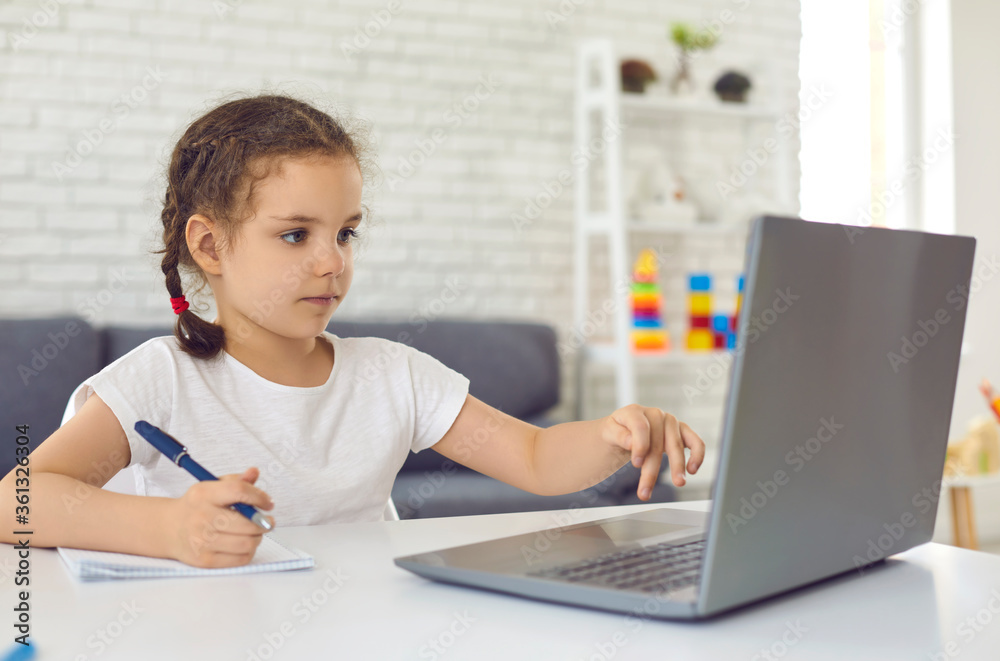 Online education. Video lesson school for children. A little girl is watching a video lesson in a laptop sitting at a table in the room. Online education. Video lesson school for children. A little girl is watching a video lesson in a laptop sitting at a table in the room.