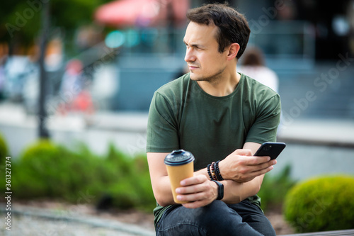 Cheerful young man using mobile phone while sitting at the city park, drinking coffee