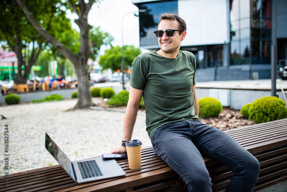 Obraz premium Handsome young man dressed casually spending time outdoors at the city, using laptop computer while sitting on a bench and drinking takeaway coffee