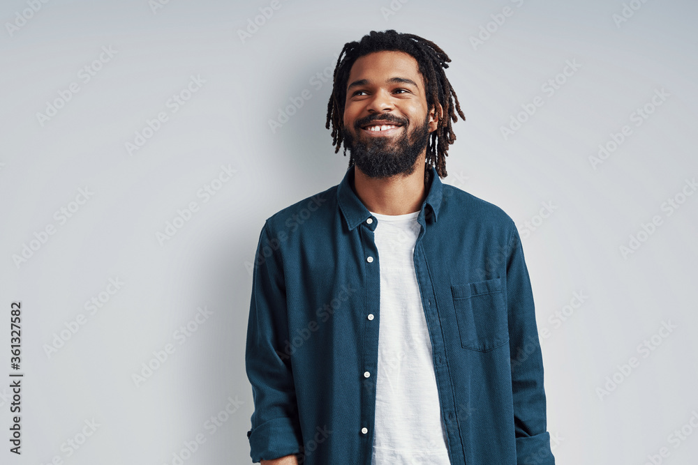 Charming young African man in casual wear smiling and looking away while standing against grey background Charming young African man in casual wear smiling and looking away while standing against grey background