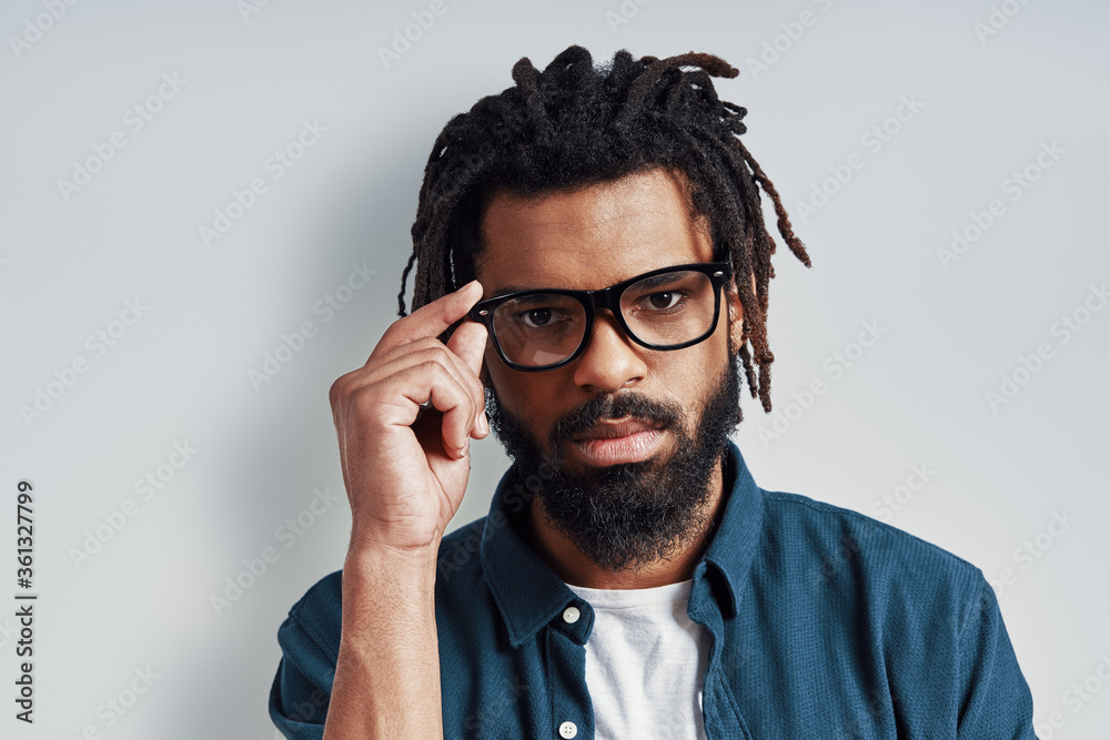 Charming young African man in casual wear looking at camera and adjusting eyewear while standing against grey background Charming young African man in casual wear looking at camera and adjusting eyewear while standing against grey background