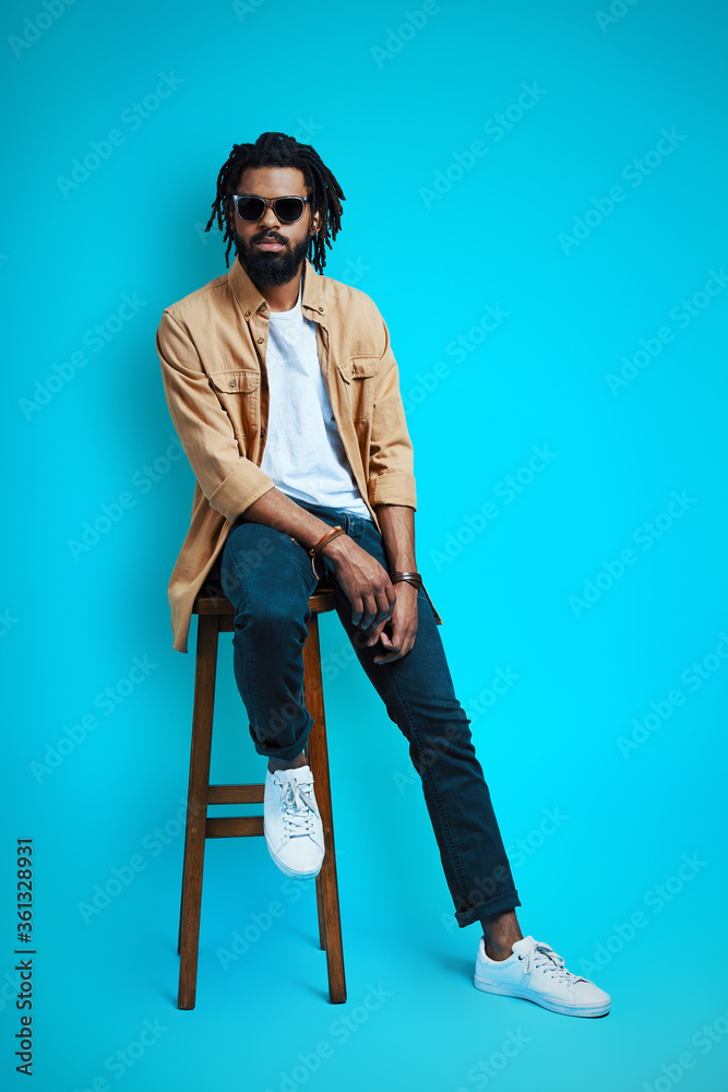Full length of charming young African man in casual wear looking at camera while sitting on stool against blue background Full length of charming young African man in casual wear looking at camera while sitting on stool against blue background