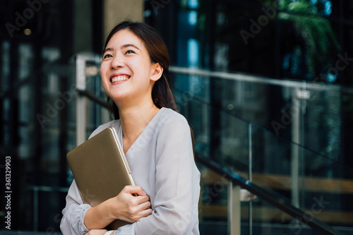 One successful and happy young adult business woman holding a laptop with smile. Professional female worker standing in corporate modern office and window in casual gray look. - With copy space.