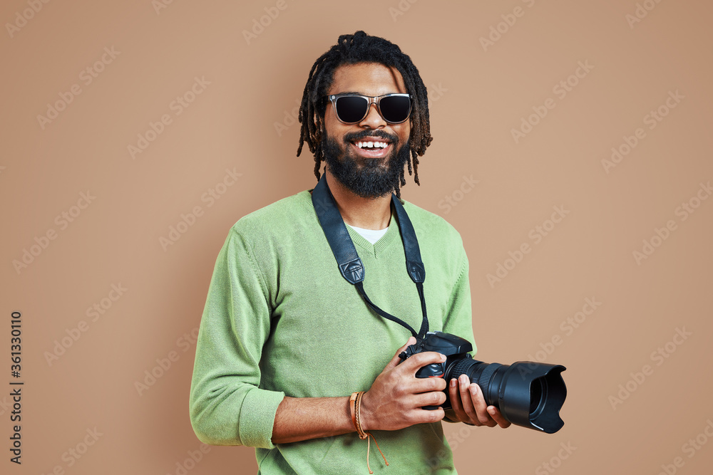 Young African photographer in casual clothing looking at camera and smiling while standing against brown background Young African photographer in casual clothing looking at camera and smiling while standing against brown background