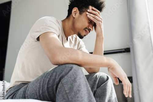 Photo of unhappy african american man with headache sitting on bed
