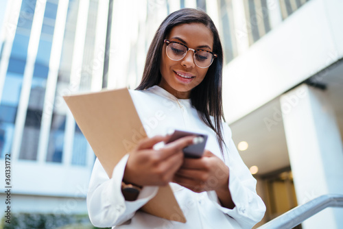 Below view of successful dark skinned employee happy with received good news in message on smartphone.Cheerful african american female lawyer with folder chatting online on cellular walking on street Free Download Below view of successful dark skinned employee happy with received good news in message on smartphone.Cheerful african american female lawyer with folder chatting online on cellular walking on street - GFXLoad