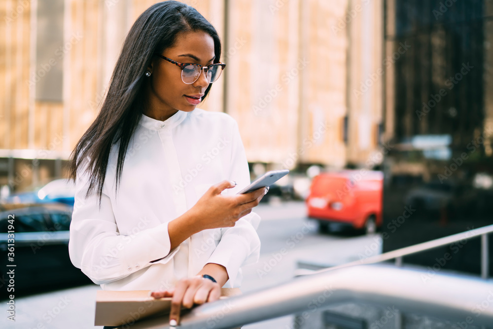 Pensive stylish young woman with dark skinned reading incoming sms on smartphone standing in downtown.African american office worker checking banking account online on cellular via 4G internet
