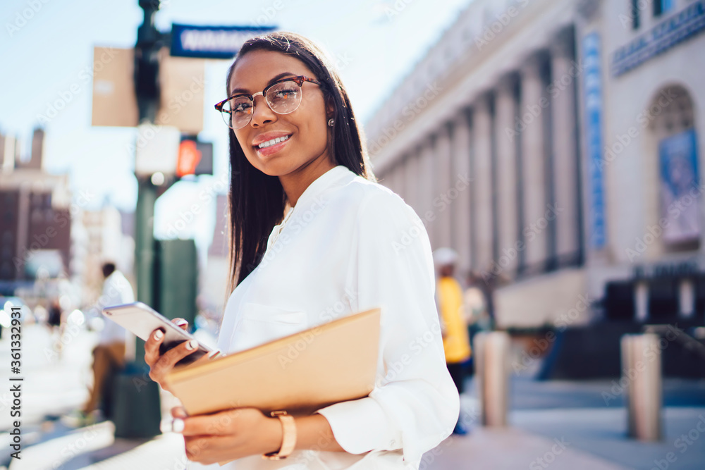 Portrait of prosperous african american businesswoman in white shirt strolling in downtown with smartphone in hands.Cheerful student of faculty of law with telephone and folder smiling at camera