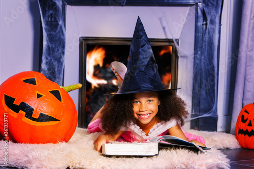 Witch girl sit with magic stick wizard hat and spell book by fireplace - Halloween portrait