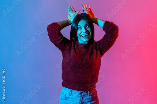 Happiness, carefree mood. Neon light portrait of joyful beautiful woman behaving childish humorous with bunny ears gesture on head, entertaining and fooling around. indoor studio shot isolated