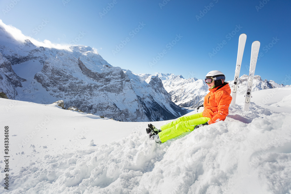 Happy girl in orange bright sport outfit with ski over high mountain range peaks and blue sky