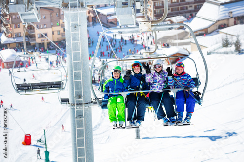 Four friends children sit together on chairlift lifting on the mountain with crowded slope and station below