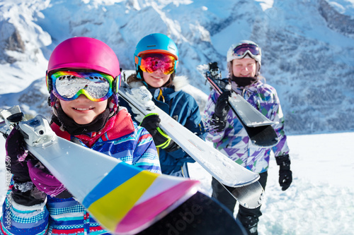 Portrait of a cute girl hold ski on shoulder with group of kids friends in colorful outfit masks and helmets in the mountains © Sergey Novikov