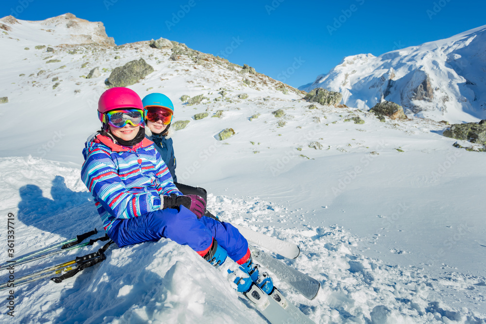 Two happy girls sit on the snow in ski masks and helmets looking at the mountains