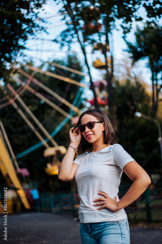 a girl with dark glasses takes off and puts them on the background of the Ferris wheel in the park. girl in a white T-shirt, and the Ferris wheel of different colors