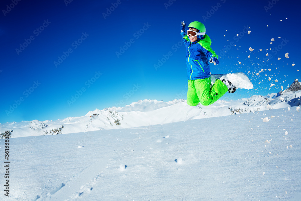 Happy boy jump on snowboard over blue sky and mountain top lifting hands with snow powder in the air