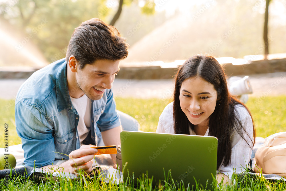 Image of couple using laptop and holding credit card while lying on grass Image of couple using laptop and holding credit card while lying on grass