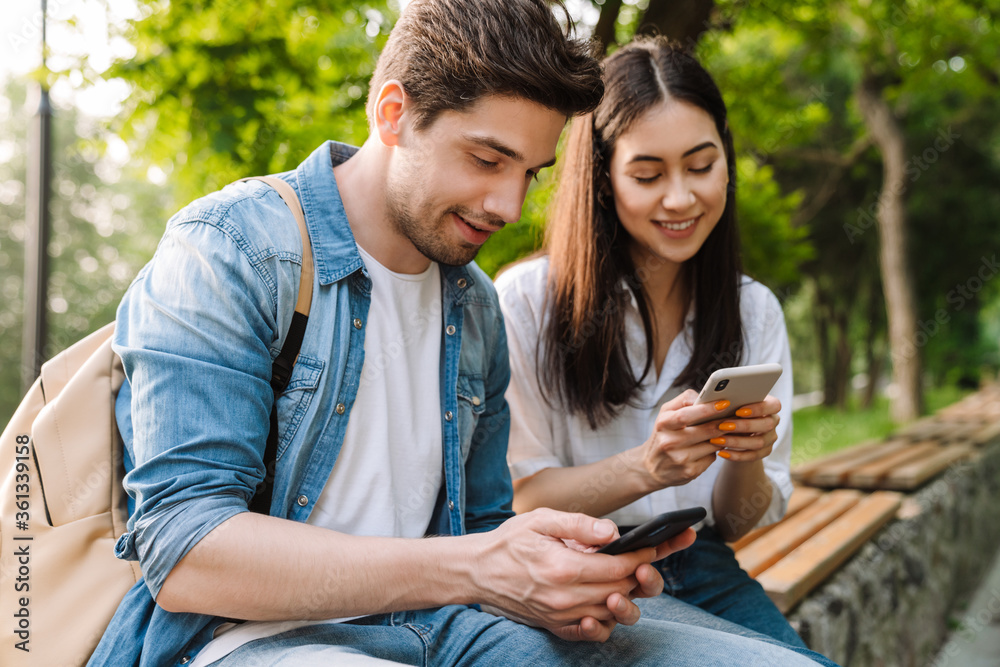 Image of pleased couple using mobile phones while sitting on bench