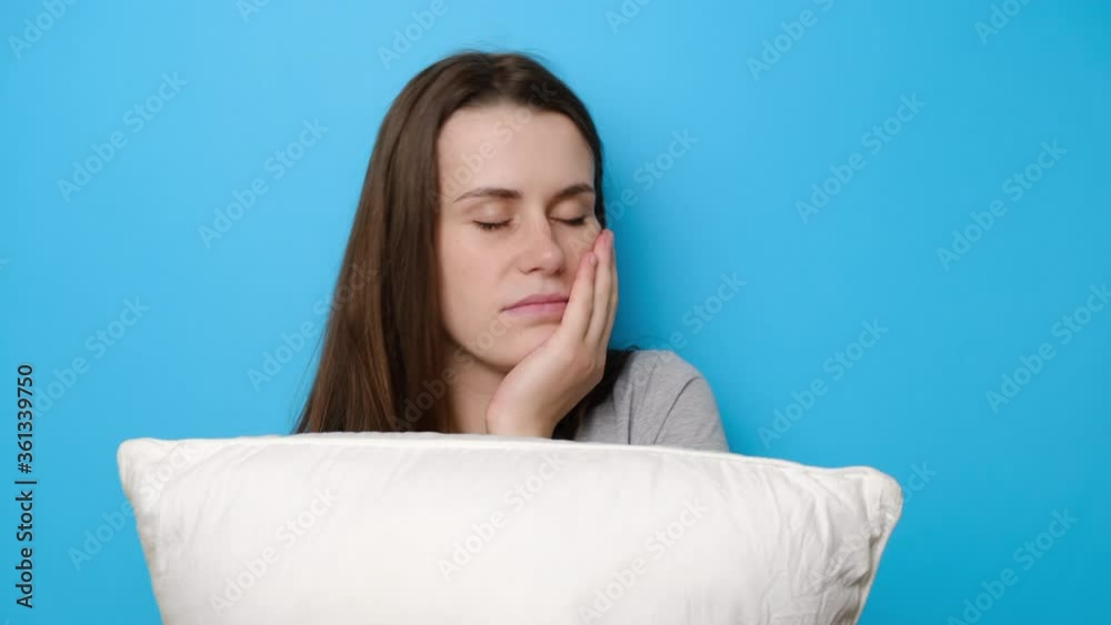 Sad dejected brunette girl holds white pillow, looks tired, suffers from insomnia, has problems with sleeping, stressed because of too early wakeup, keeps hand under chin, isolated on blue background Sad dejected brunette girl holds white pillow, looks tired, suffers from insomnia, has problems with sleeping, stressed because of too early wakeup, keeps hand under chin, isolated on blue background